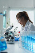 © crizzystudio - Young female scientist wearing lab coat and protective glasses using microscope while analyzing samples in modern laboratory, surrounded by test tubes filled with blue liquid
