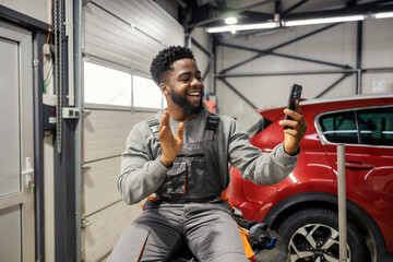 Poster - Smiling black repairman sitting on car tires at auto mechanic workshop and waving at the phone during video call.