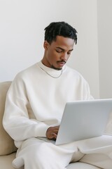 Wall Mural - A man wearing a white shirt and pants is sitting on a couch and using a laptop. He is focused on his work or browsing the internet