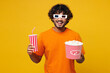 © ViDi Studio - Young smiling happy Indian man he wear orange t-shirt casual clothes 3d glasses watch movie film hold bucket of popcorn cup of soda pop in cinema isolated on plain yellow background studio portrait.