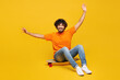 © ViDi Studio - Full body young fun Indian man he wear orange t-shirt casual clothes sitting on skateboard pennyboard raise up hands look camera isolated on plain yellow background studio portrait. Lifestyle concept.