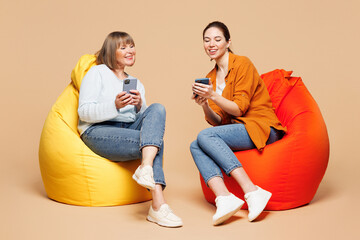 Full body elder parent mom 50s years old with young daughter two women together wear casual clothes sit in bag chair use show mobile cell phone isolated on plain beige background. Family day concept.