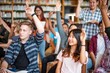© Rawpixel.com - Diverse group of students in a library raising hands. Engaged students, mixed ethnicities, actively participating in a learning environment. Diverse students in library, education concept.