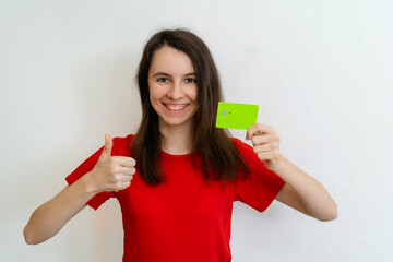 smiling young brunette woman wears basic red t-shirt hold in hand credit bank card show thumb up ges