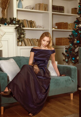 Beautiful young blonde in an evening dress sits on a sofa against the background of a Christmas tree