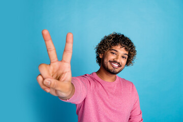 Wall Mural - Smiling young man making a peace gesture with hand against a blue backdrop, embodying happiness and modern casual fashion in a pink sweater