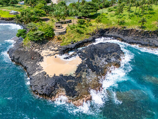 Canvas Print - Boca de Inferno, Sao Tome, featuring black volcanic rock formations, turquoise ocean waves, and a lush green hillside covered with tall, swaying palm trees under a clear blue sky.