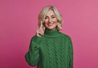 Poster - Portrait of a beautiful, smiling middle-aged woman with blonde hair, wearing a green sweater and pointing her finger downward, isolated on a pink background