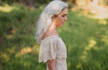 A stylish woman with flowing white hair stands in a lush green field, captured in natural sunlight. Her lace dress and serene expression convey elegance and tranquility in a pastoral setting.