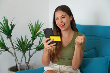 Canvas Print - Happy young Brazilian woman holding smartphone and credit card celebrates with a clenched fist sitting on couch at home