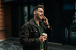 © dikushin - Portrait of cheerful young man enjoying coffee from disposable cup while talking on mobile phone, walking relaxing along city street. Concept of modern communication and urban lifestyle.