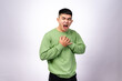 © Daniel - An Asian man in a green sweatshirt clutches his chest with both hands, his face showing discomfort and pain. He appears to be experiencing chest pain or distress. The background is plain white