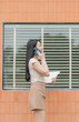 © Kritdanai - Modern Communication: A professional woman in a beige ensemble engaging in a phone conversation, holding a tablet in front of a window, creating an image of contemporary connectivity and work life.