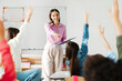 © Home-stock - Friendly happy teacher asking pupils while kids raising hands want to ask questions or answer, sitting at their desks in classroom