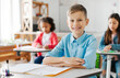 © Home-stock - Portrait of positive schoolboy sitting at school desk looking and smiling at camera during lesson, classmates on background