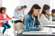 © Home-stock - Smart schoolgirl sitting at desk and writing in copybook, doing task creating studious atmosphere in classroom interior, private school
