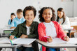 © Home-stock - Happy black friends schoolboy and schoolgirl smiling at camera, sitting at desks in classroom with other children on background