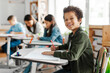 © Home-stock - Happy Latin schoolboy sitting at desk and smiling at camera, classmates writing on the background, pupils creating studious classroom atmosphere