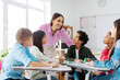 © Home-stock - Group of diverse kids listening their friendly female teacher while sitting at desks in classroom, creating positive atmosphere