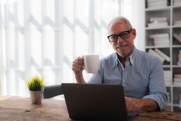 Wall Mural - Happy businessman enjoying a coffee break while working on a laptop in a modern office, smiling and embracing a moment of relaxation during a busy day