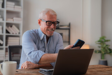 Happy mature businessman in blue shirt sitting at desk in modern office, holding mobile phone and comparing data between laptop and smartphone, smiling