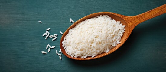 Poster - White jasmine rice heaped in a wooden spoon on a dark teal background with scattered grains in the foreground, showcasing a natural, rustic feel