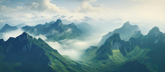 Aufkleber - Aerial view of lush green mountains shrouded in mist under a soft blue sky with scattered clouds, capturing a serene and majestic landscape.