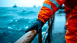 © FTN-STUDIO - A worker in an orange raincoat grips a railing on a boat, with turbulent blue waters in the background, illustrating maritime labor in challenging conditions.