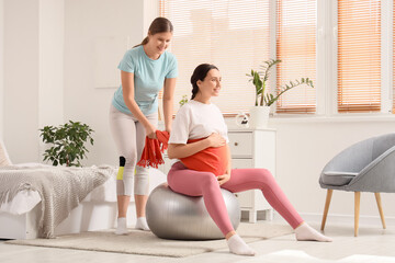 Wall Mural - Young doula with scarf massaging pregnant woman on fitball in bedroom