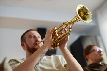 Wall Mural - Close up of red haired man playing trumpet enthusiastically while sitting on couch at home with friends, copy space