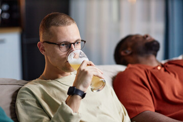 Wall Mural - Portrait of adult man wearing glasses and drinking beer from tall glass while watching TV on couch with friends, copy space