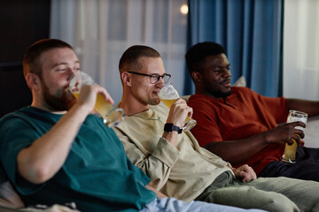 Wall Mural - Group of three friends watching TV together sitting on couch and drinking beer in blue tv light, copy space