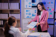 © Tj - Two Asian businesswomen collaborating on a project, exchanging documents filled with financial charts and graphs in a modern office environment, exemplifying teamwork and professionalism