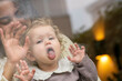 © Connect Images - Baby with curly hair pressing face against glass, mother in background smiling and waving. Netherlands