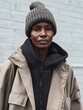 © Jaemie - Urban Fashion: A model wearing a black hoodie and beige oversized coat, topped with a gray knit beanie against a muted backdrop.