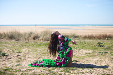 Wall Mural - Woman dancing flamenco in a white dress with floral print and ruffles, sitting on an old wooden chair with her head back and her hair blowing in the wind. On the horizon the Atlantic ocean.