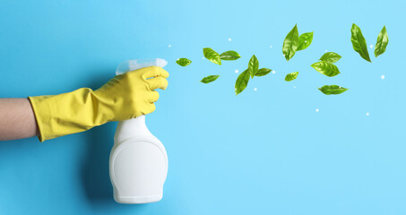 Wall Mural - Woman spraying cleaning product on light blue background, closeup. Green leaves flying out of bottle