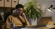 © m-art - Young curly-haired dark-haired man wearing glasses with a serious look talking on a mobile phone, angry and cursing, sitting in front of an open laptop in a modern stylish office