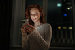 © dikushin - Portrait of happy young woman browsing social media on smartphone, enjoying cozy evening at home, soft screen light illuminating smiling face in tranquil bedroom atmosphere on background of window.
