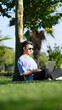 © HI Pictures - A businessman working happily on his laptop while leaning against a tree trunk in a tree-filled park on a summer day