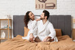 © Pixel-Shot - Happy couple in bathrobes sitting on bed at home