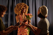 © pressmaster - Woman with curly hair, wearing a colorful dress, smiling while receiving flowers from two other women in front of draped curtains