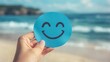 © Arfi Studio - Hand holding a blue circle with a smiling face on a beach.