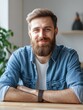 © Johannes - Smiling young adult bearded casual man looking at camera posing alone at home office workplace. Millennial guy student, freelancer sitting at table satisifed with distance remote job concept. Portrai