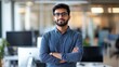 © Sirat - Confident young professional man standing with arms crossed in modern office environment with computers and natural light in background