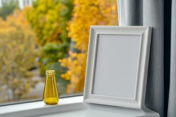 Poster - A close-up photo of an empty white picture frame on top of the window sill in front of large windows with light gray curtains. Next to it is a lamp and a bottle. The background outside shows