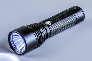 Poster - A small flashlight is lying on the table, illuminated by its beam of light. The flashlight has an elongated shape with a textured metal body and a black matte finish. It features multiple buttons