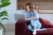 © ingoodtime - Modern freelancer woman working remotely from home, multitasking with laptop and smartphone while sitting on cozy sofa. Focused tech-savvy female manages online business tasks on comfortable workplace