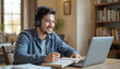 © aiartth - A positive Hispanic student man wearing headphones watches an online lecture while taking notes in a copybook, sitting at a home study table with open books.