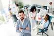 © deagreez - Confident businessman stands in a busy office surrounded by coworkers collaborating during a team meeting